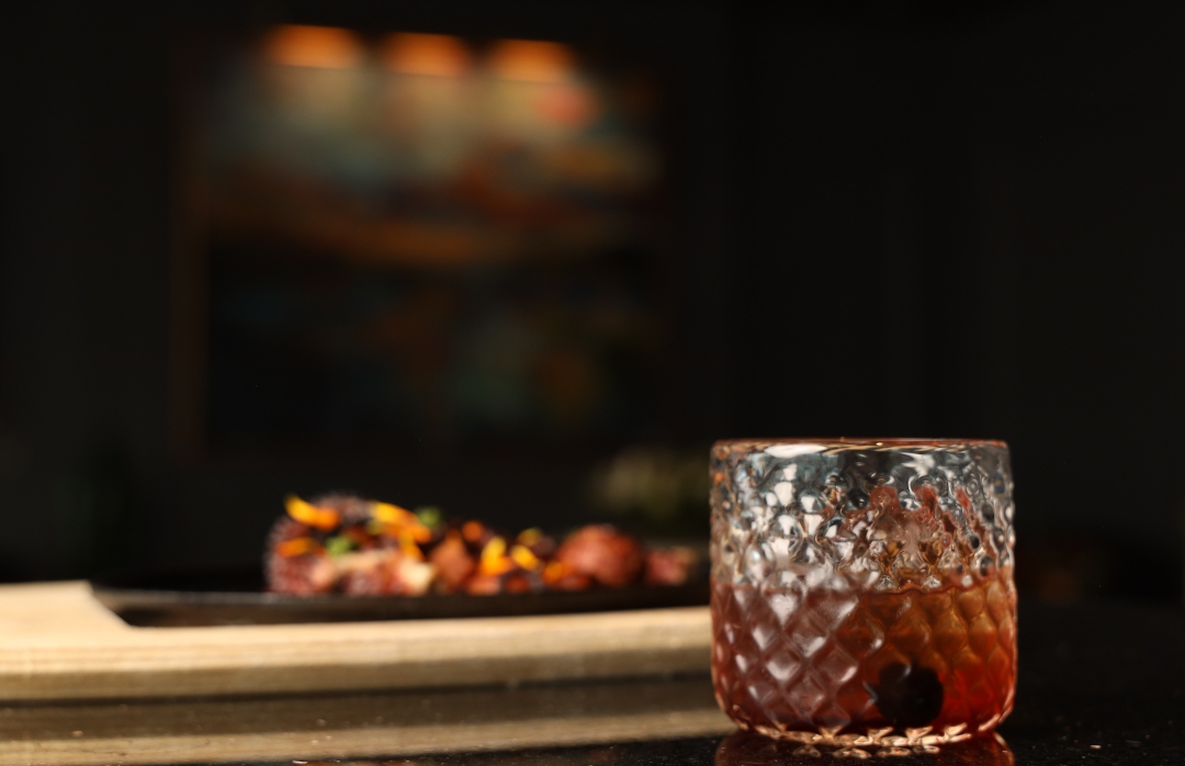 A close-up shot of a textured, patterned rocks glass containing an amber cocktail with a dark cherry garnish.