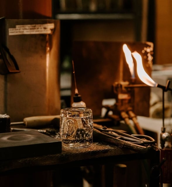 A clear, patterned glass tumbler with embedded bubbles rests on a dark workbench. Tools and a bright torch flame are visible in the warm-lit workshop background.
