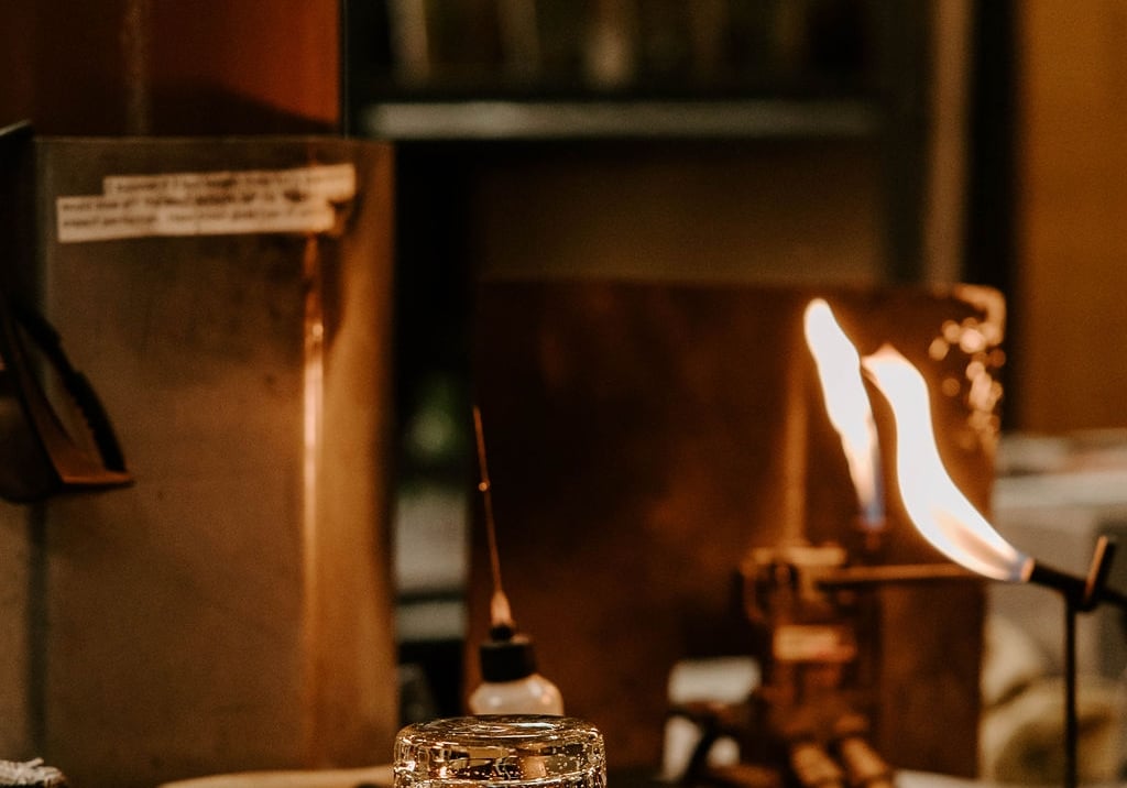 A small glass cup sits on a workshop table next to tools and an active torch flame in a glassblowing or craft studio.