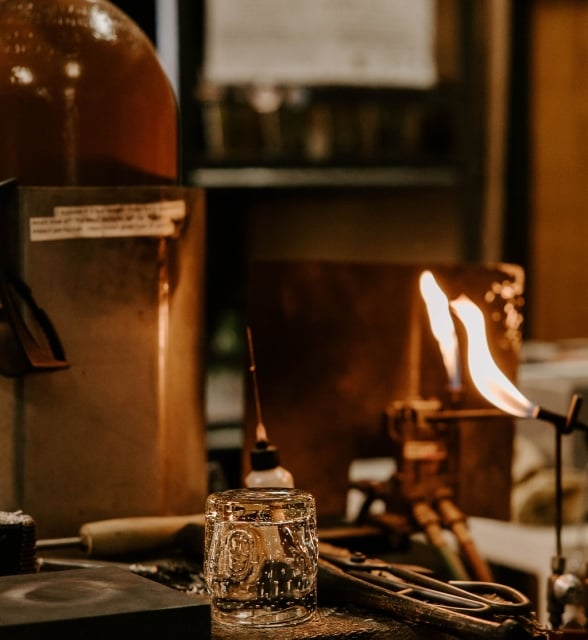 A small glass cup sits on a workshop table next to tools and an active torch flame in a glassblowing or craft studio.