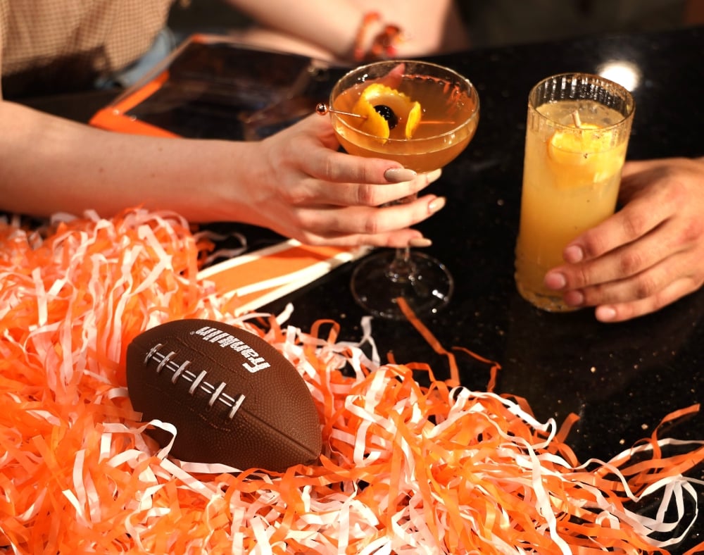 A close-up shot of a dark table setting featuring two cocktails, an orange and white shaker, and a small football resting on a bed of orange and white paper shreds.