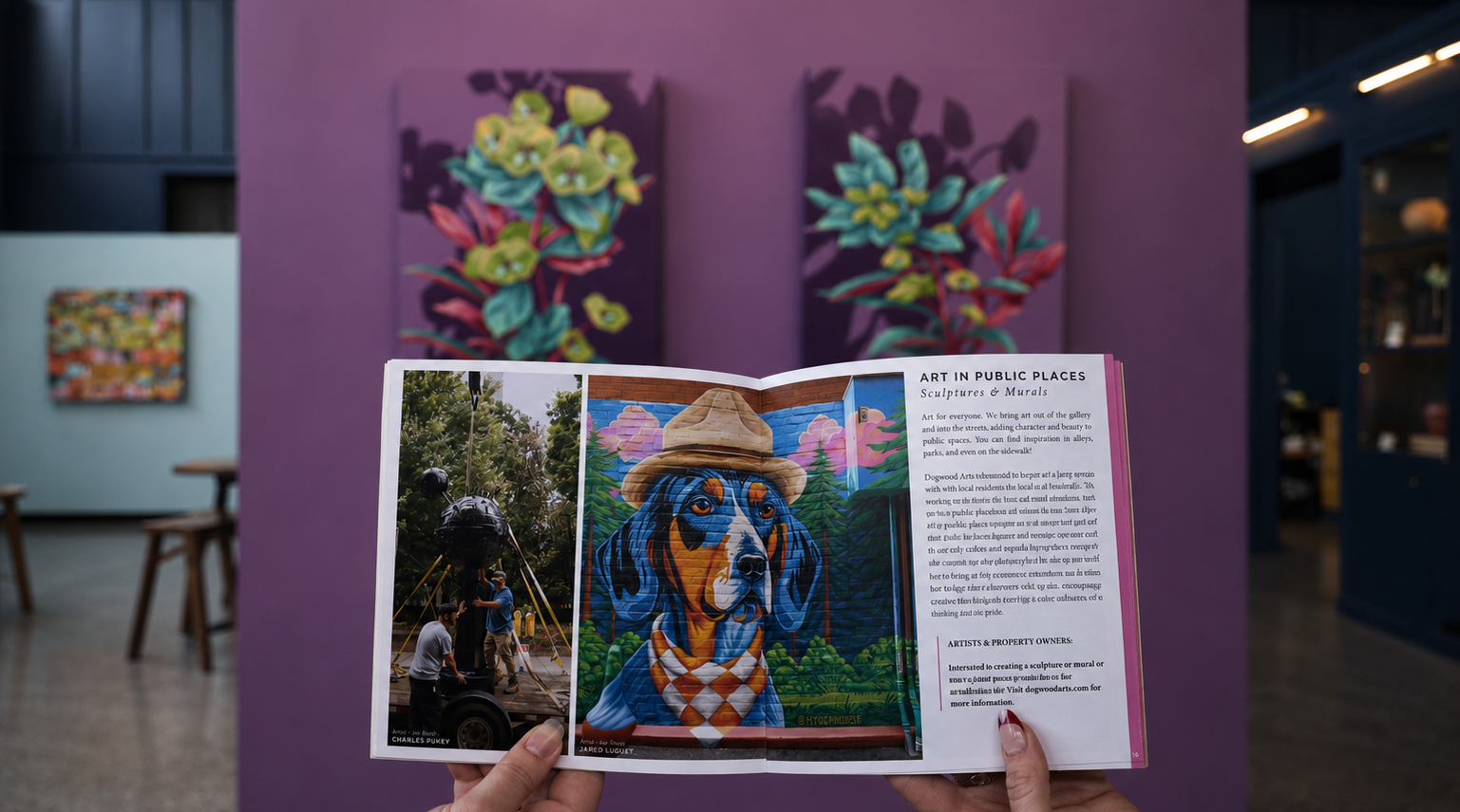 Hands hold an open booklet showcasing public art projects, including a dog mural and a sculpture installation, against a blurred art gallery. Text: "ART IN PUBLIC PLACES Sculptures & Murals".