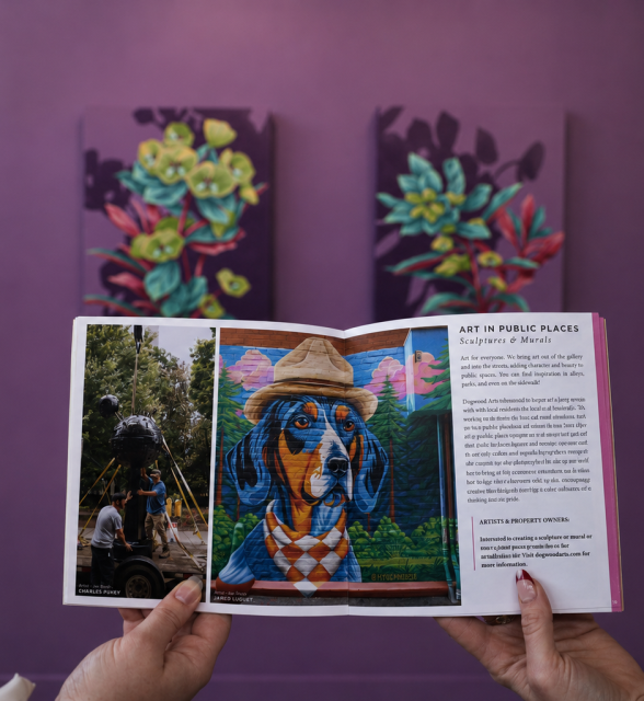 Hands hold an open booklet showcasing public art projects, including a dog mural and a sculpture installation, against a blurred art gallery. Text: "ART IN PUBLIC PLACES Sculptures & Murals".