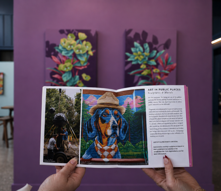 Hands hold an open booklet showcasing public art projects, including a dog mural and a sculpture installation, against a blurred art gallery. Text: "ART IN PUBLIC PLACES Sculptures & Murals".
