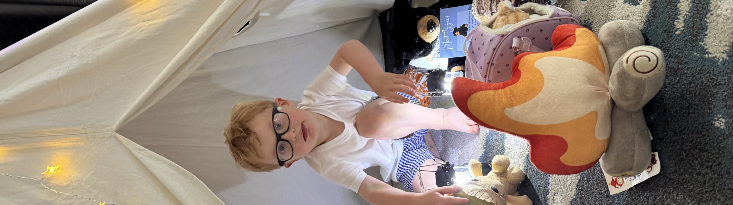 A young boy with glasses sitting inside a white teepee play tent decorated with star-shaped fairy lights.