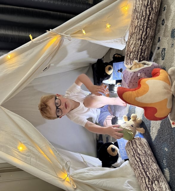 A young boy with glasses sitting inside a white teepee play tent decorated with star-shaped fairy lights.