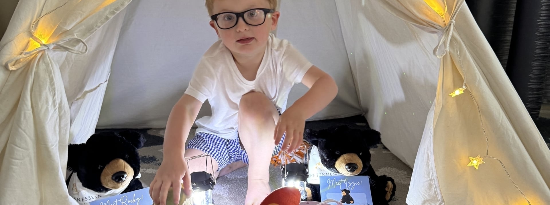 A young boy with glasses sitting inside a white teepee play tent decorated with star-shaped fairy lights.