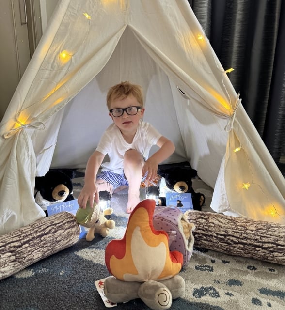 A young boy with glasses sitting inside a white teepee play tent decorated with star-shaped fairy lights.