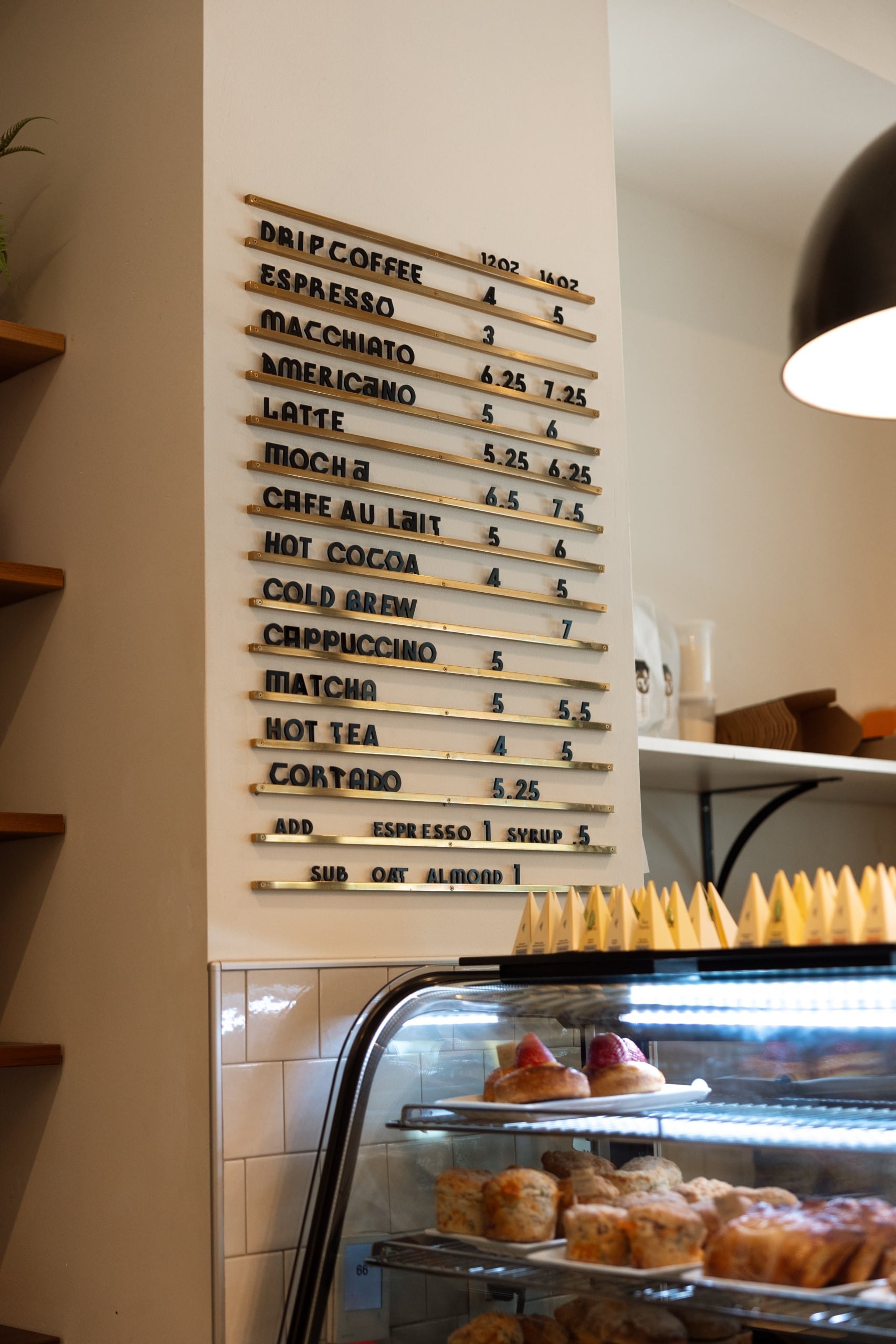 Café menu board with black letters on brass rails lists coffee (Drip Coffee 12oz 16oz, Espresso 4 5) and tea. It's above a pastry display case.