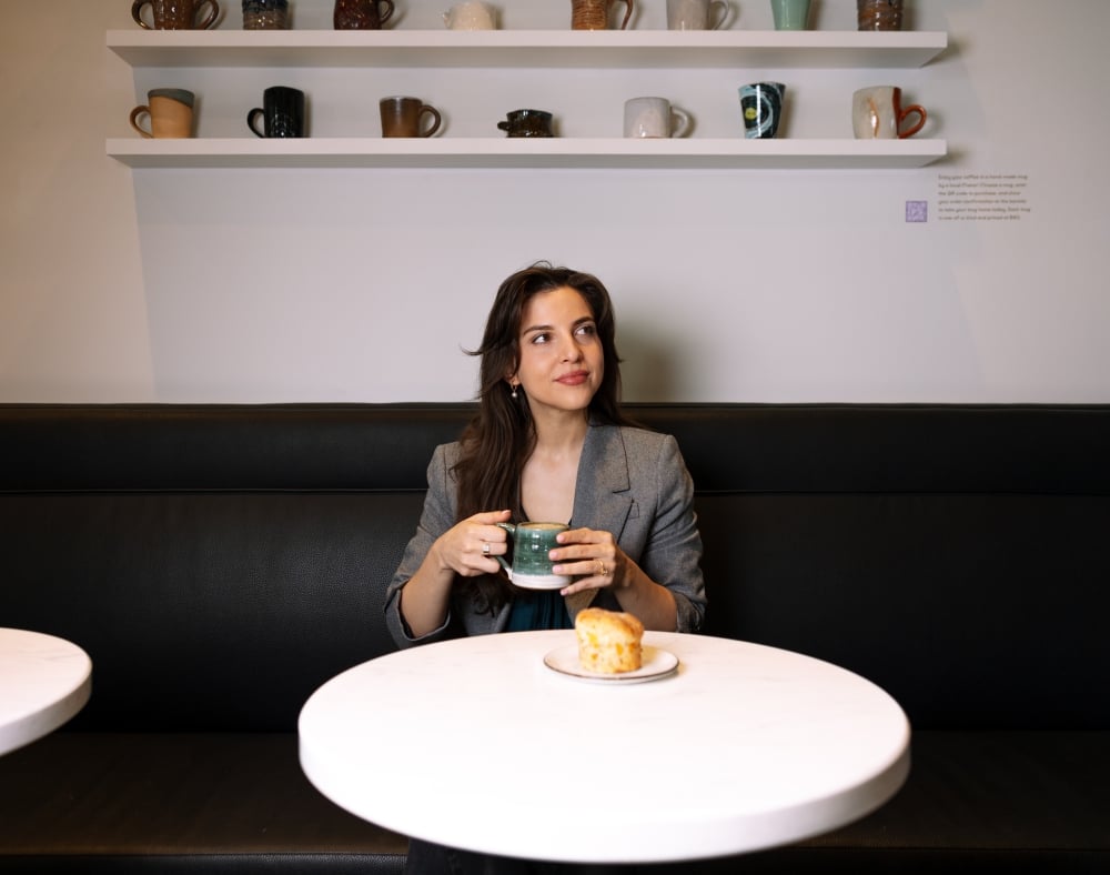 A smiling woman sitting at a white marble cafe table, looking off-camera while holding a green ceramic coffee mug.