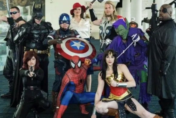 A group of cosplayers dressed as various comic book and movie characters—including Batman, Captain America, Spider-Man, Wonder Woman, Thor, and Nick Fury—posing together for a photo at Fanboy Expo.