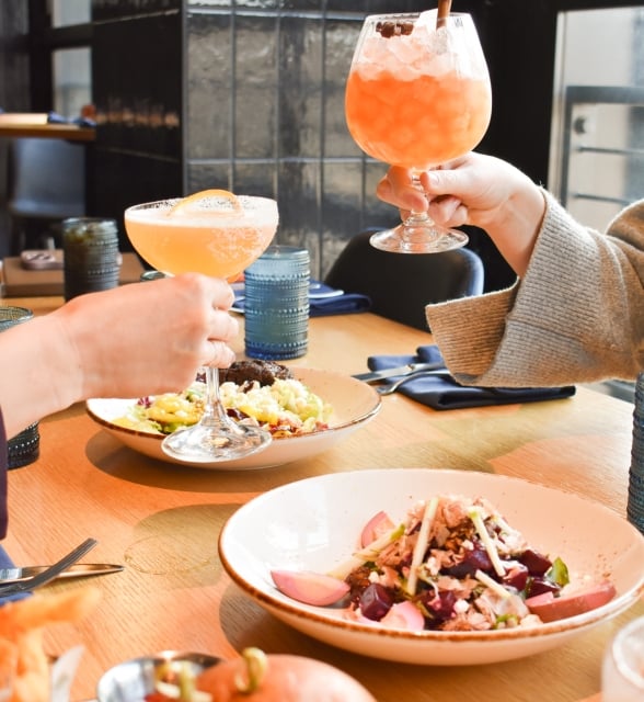 Two hands raise and clink orange cocktails above a wooden table laden with several dishes, including salads and a burger, and other drinks, in a restaurant setting.