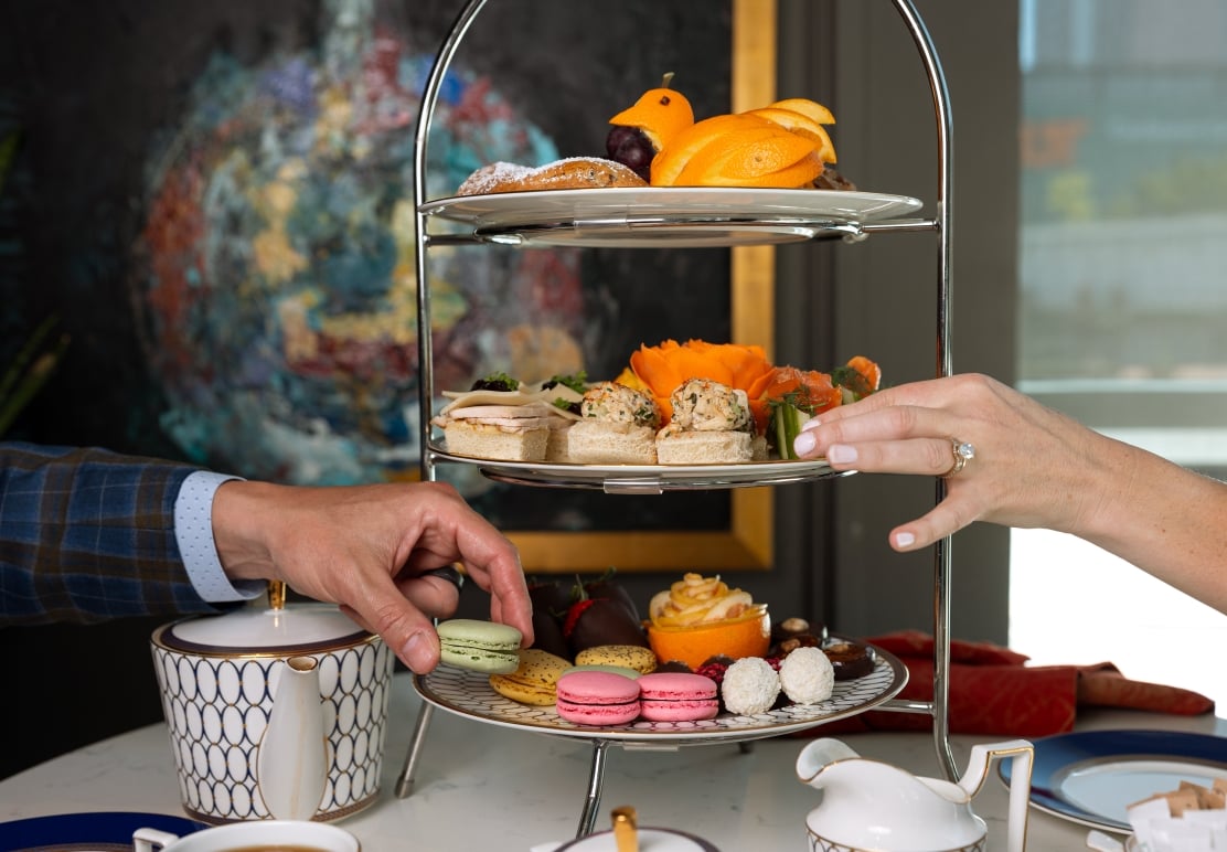 A close-up shot of a tiered dessert stand featuring sandwiches, macarons, and fruit, with two hands reaching for treats.