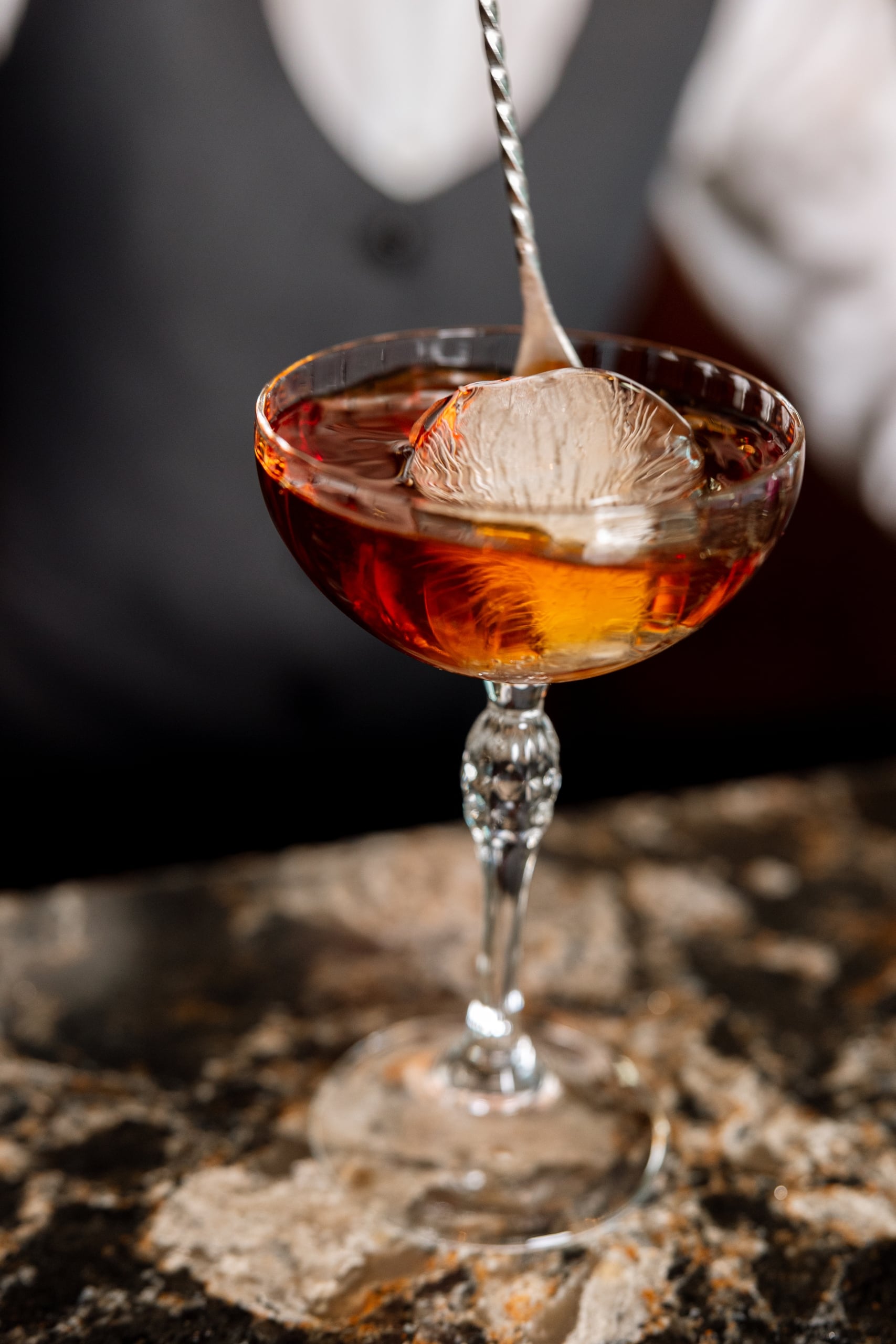 A coupe glass with an amber cocktail and a spherical ice cube is stirred by a twisted bar spoon. It rests on a speckled counter, with a blurred bartender in the background.