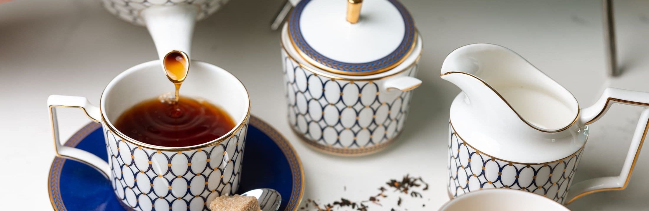 Hands pouring tea from a white and gold teapot into a blue and gold teacup on a marble table, next to a matching tea set and a three-tiered high tea tray filled with pastries, macarons, and sandwiches.