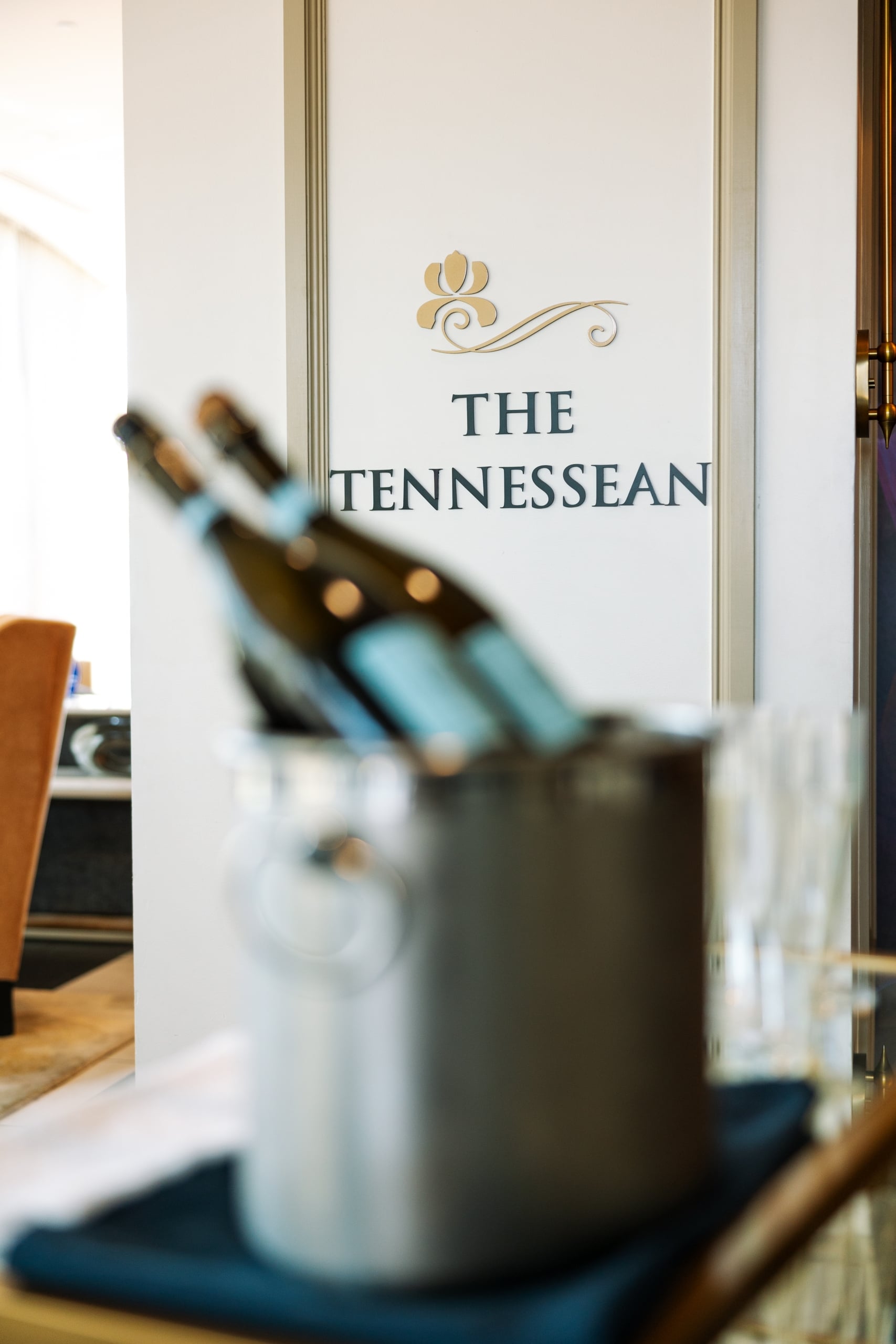Wine bottles chill in a metal bucket, blurred in the foreground, against a white wall displaying a gold floral logo and "THE TENNESSEAN".