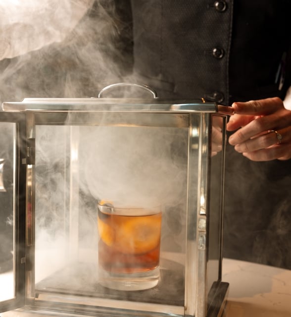 A cocktail in a glass smoking box is being opened by a person wearing a dark vest, releasing thick smoke across a white counter.