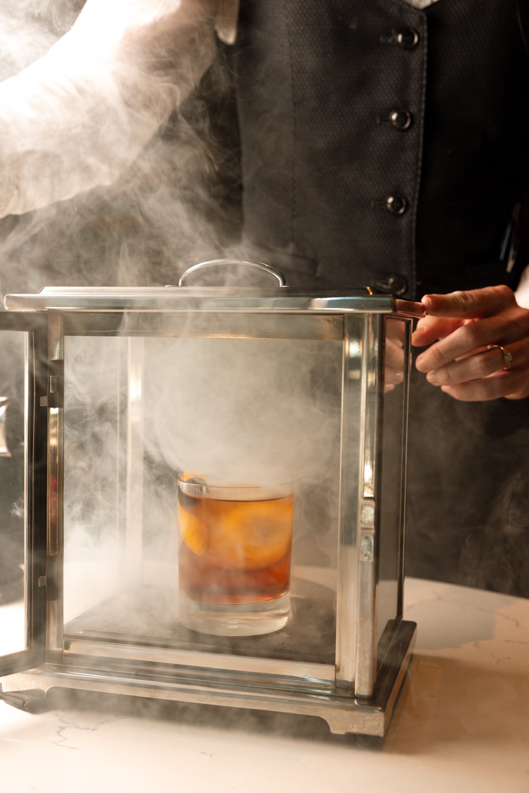 A cocktail in a glass smoking box is being opened by a person wearing a dark vest, releasing thick smoke across a white counter.