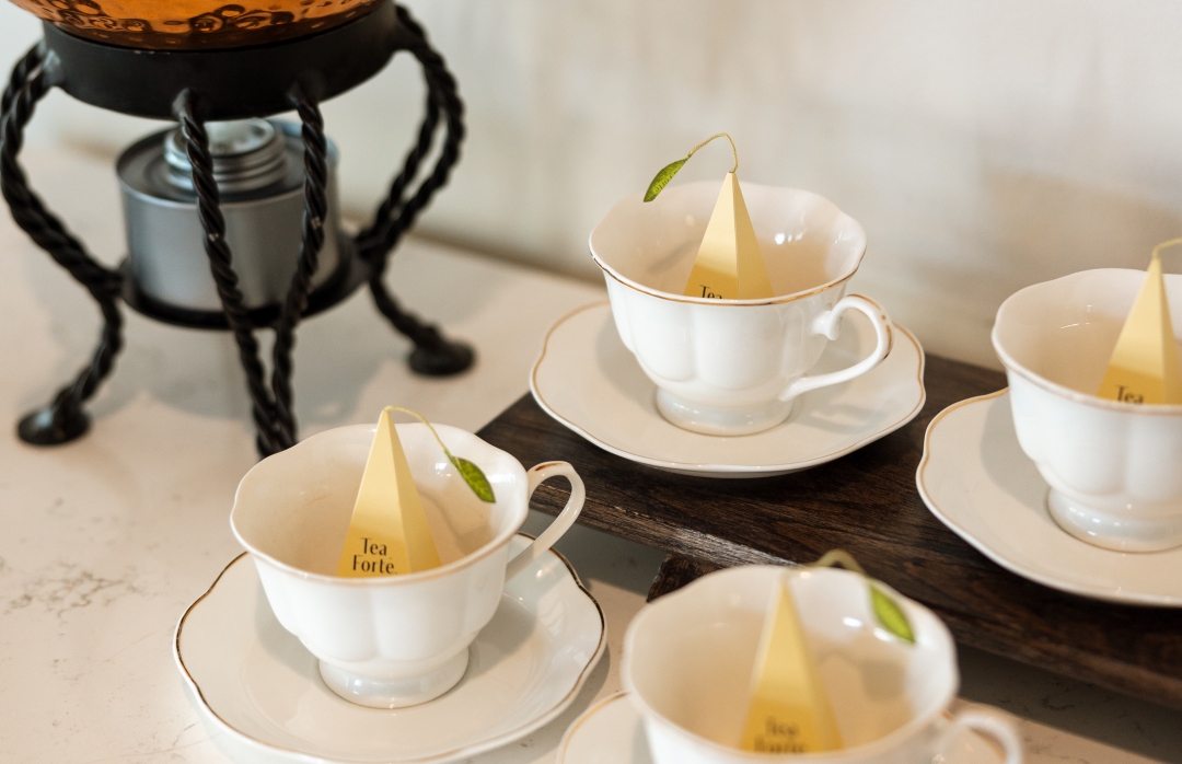 Four white porcelain teacups with Teaforté pyramid infusers inside, arranged on a tray next to a copper tea urn.