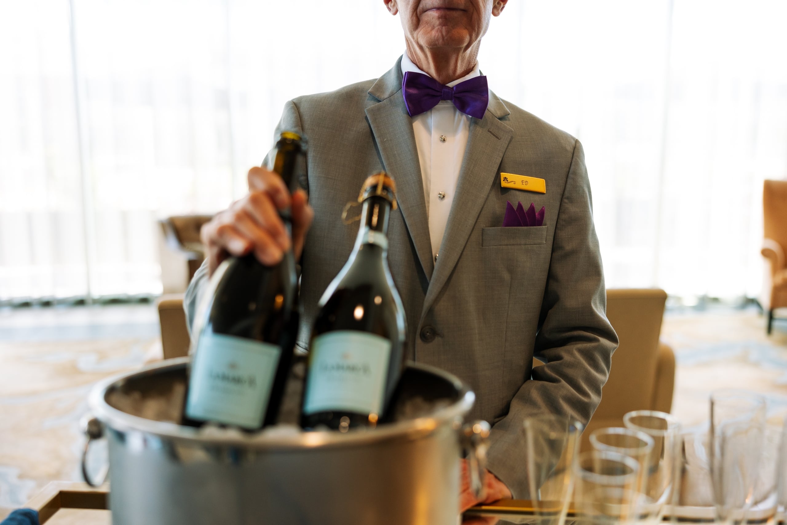 A server in a grey suit and purple bow tie holds a "LABARRAUD" sparkling wine bottle from an ice bucket. A name tag reads "ED". Bright background.