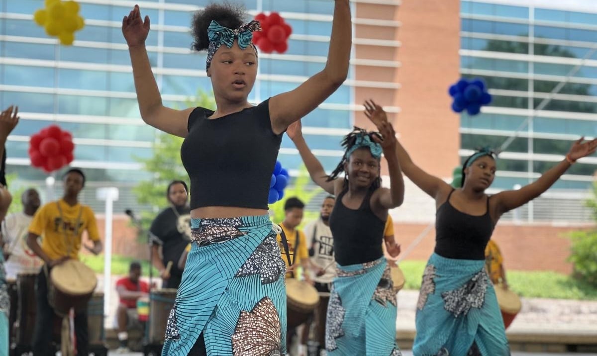 Young dancers in matching blue patterned wraps and black tops performing an outdoor routine at the Kuumba Festival, with drummers playing in the background.