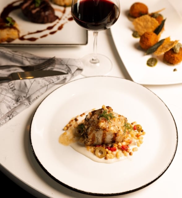 Grilled fish with sauce, vegetables, and grains is presented on a white plate. A glass of red wine and other gourmet dishes fill a white table. Napkin reads "Malones".