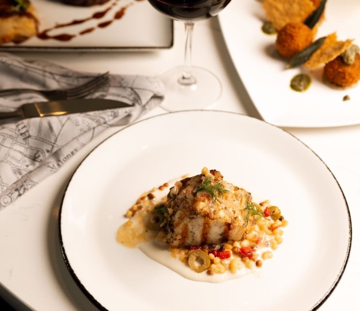 Grilled fish with sauce, vegetables, and grains is presented on a white plate. A glass of red wine and other gourmet dishes fill a white table. Napkin reads "Malones".