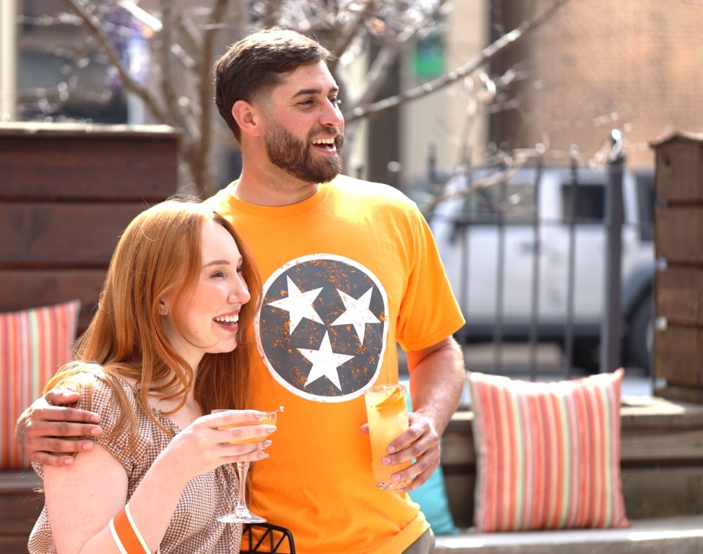 A smiling woman and a man in a Tennessee Volunteers t-shirt enjoying drinks outside on a patio.