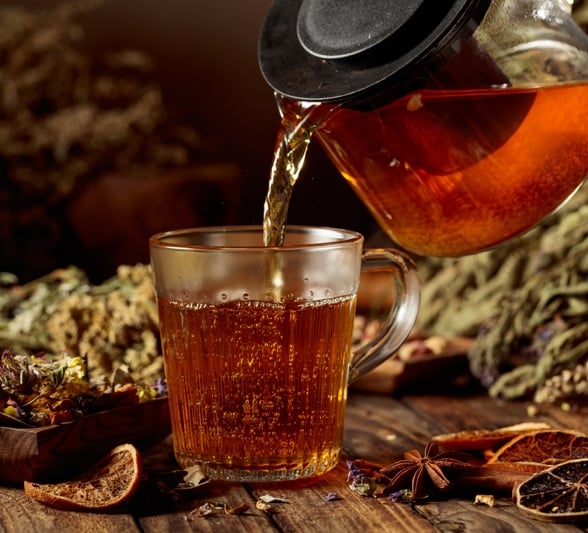 Dark amber tea pours from a glass teapot into a clear mug on a rustic wooden table surrounded by dried herbs, star anise, and orange slices.