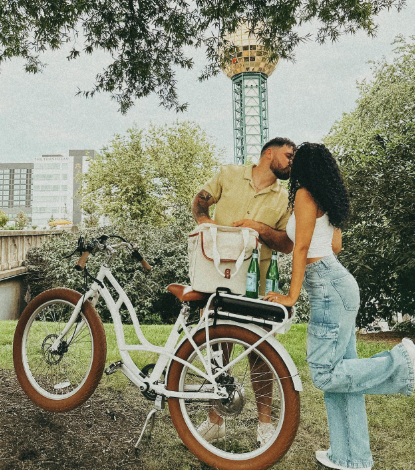 A couple kisses while leaning on a white bicycle with a cooler bag and two green bottles on its rear rack, set outdoors with trees and the Sunsphere in the background.