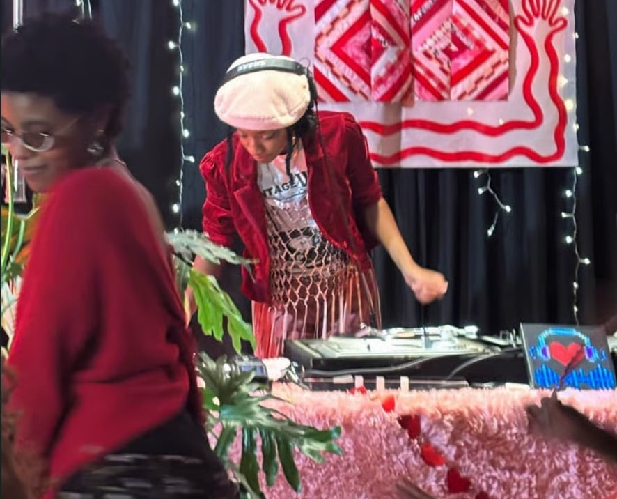Two people stand at a decorated table with a turntable, plants, string lights, and a red-and-white patterned backdrop.