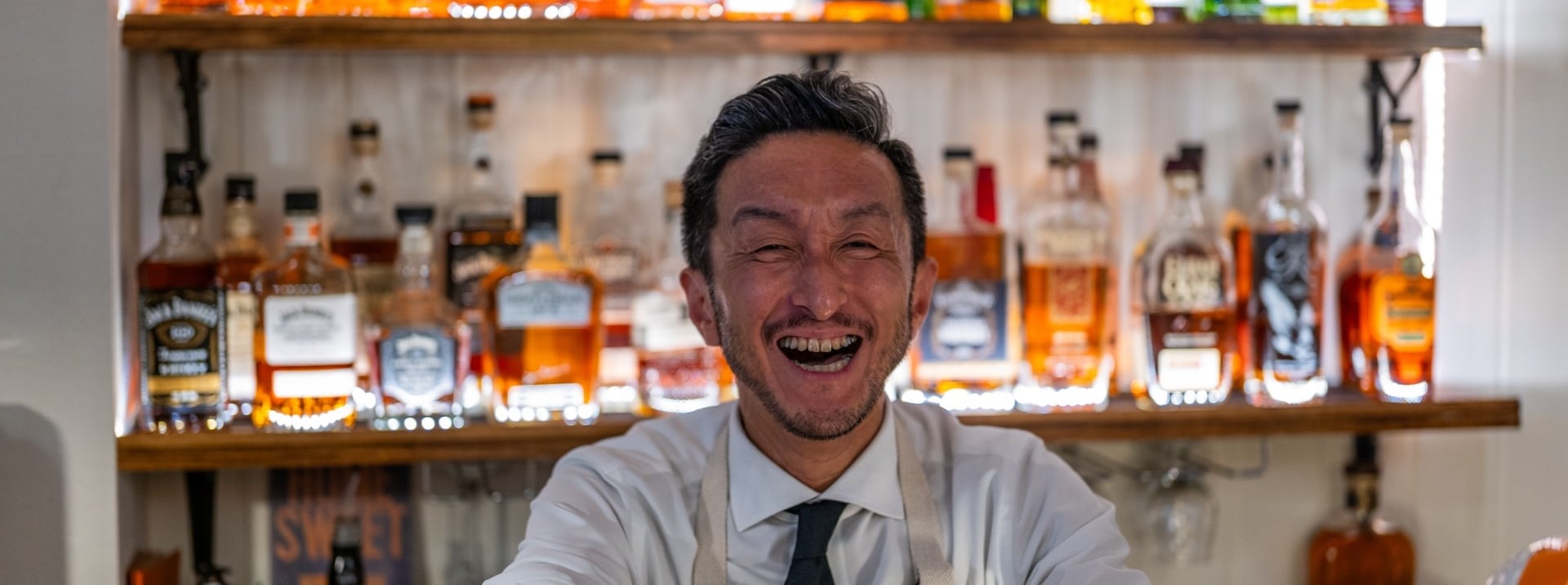 A smiling bartender in a white shirt, tie, and apron stands behind a bar, with shelves of liquor bottles behind him.