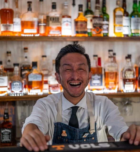 A smiling bartender in a white shirt, tie, and apron stands behind a bar, with shelves of liquor bottles behind him.