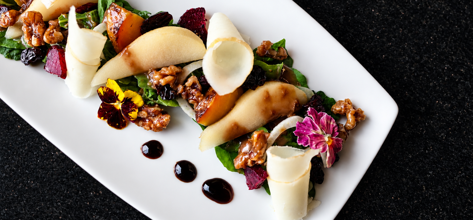 Elegant pear and walnut salad with greens, shaved cheese curls and edible flowers on a white platter with balsamic dots.