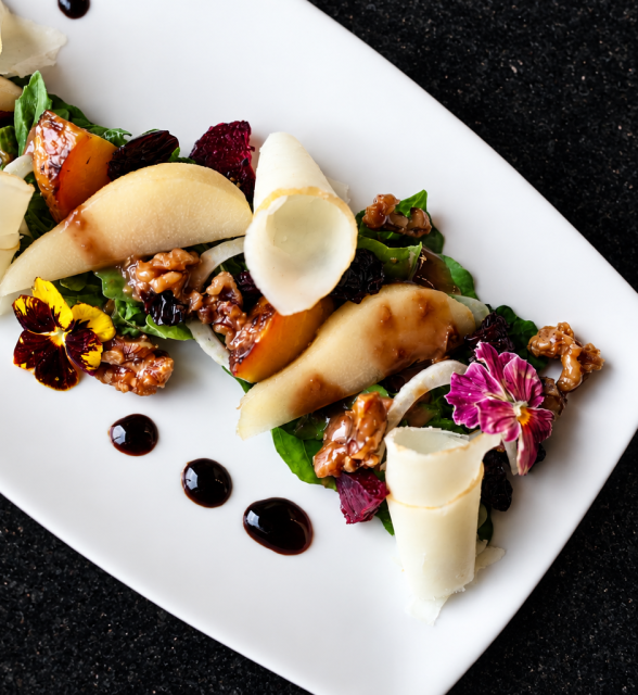 Elegant pear and walnut salad with greens, shaved cheese curls and edible flowers on a white platter with balsamic dots.
