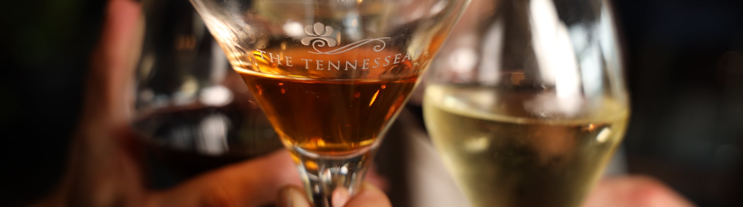 Close-up of hands clinking cocktail glasses during a toast, with one glass etched reading 'THE TENNESSEE'.
