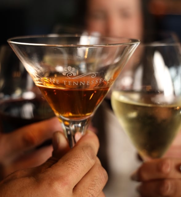 Close-up of hands clinking cocktail glasses during a toast, with one glass etched reading 'THE TENNESSEE'.