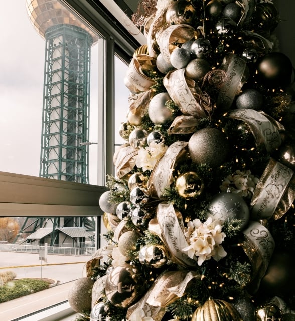 Tall Christmas tree decorated with gold and silver ornaments, ribbons, and white flowers by a window.